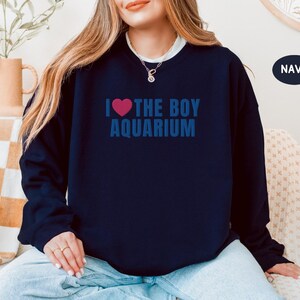 May include: Navy blue sweatshirt with the text "I ❤ THE BOY AQUARIUM" in blue, with a pink heart. The sweatshirt is a crew neck style. The word "NAVY" is in a navy blue circle in the upper right corner.