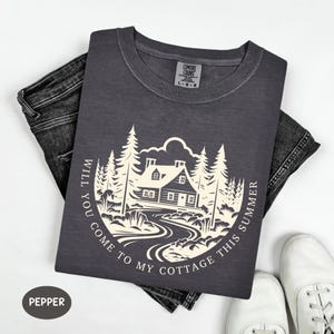 May include: A dark gray t-shirt with a cream-colored graphic of a cabin in a forest, with the text "WILL YOU COME TO MY COTTAGE THIS SUMMER" around the image. The shirt is paired with dark jeans and white sneakers.