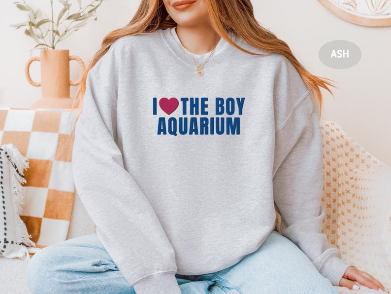 May include: Light gray sweatshirt with the text "I ❤️ THE BOY AQUARIUM" in navy blue and a red heart. The sweatshirt is a casual style, suitable for everyday wear.