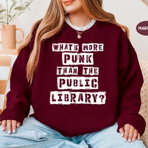 Könnte beinhalten: Burgunderfarbenes Sweatshirt mit dem weißen Text "WHAT'S MORE PUNK THAN THE PUBLIC LIBRARY?". Das Sweatshirt hat einen Rundhalsausschnitt und lange Ärmel. Das Wort "MAROON" steht in einem weißen Feld oben rechts.