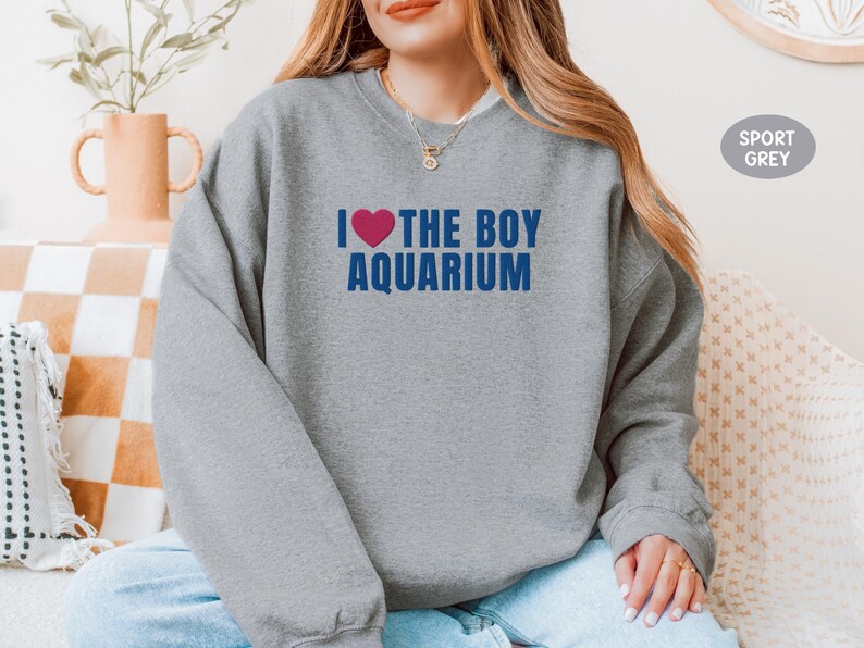 May include: Sport grey sweatshirt with the text "I ❤️ THE BOY AQUARIUM" in blue and a pink heart. The sweatshirt is a casual style, perfect for everyday wear. The text is centered on the chest.