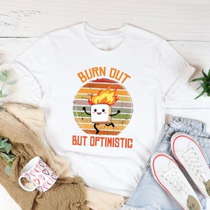 May include: White t-shirt with a retro-style graphic of a burning marshmallow with a smiling face. The text "Burn Out But Optimistic" is printed below the graphic.