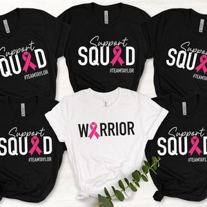 May include: Five black t-shirts with white text and a pink ribbon graphic. The text on four of the shirts reads "Support Squad #TEAMTAYLOR". The text on the fifth shirt reads "WARRIOR".