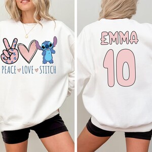 May include: White sweatshirt with a peace sign, heart, and Stitch character on the front. The back of the sweatshirt has the name "Emma" and the number "10" in pink.