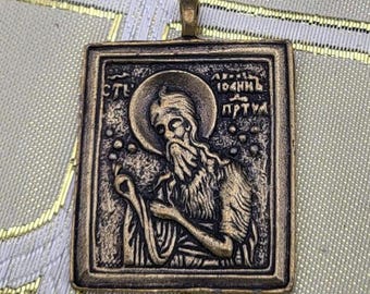 May include: A rectangular bronze pendant featuring a religious icon. The icon depicts a bearded figure with a halo, holding an object. The pendant has a loop at the top for a chain. The background is a patterned fabric.