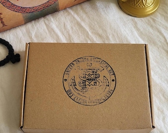 Puede incluir: Una caja rectangular de cartón marrón con un sello circular en tinta negra. El sello dice "Sacred Harbor Liturgical Art" y "sacredharborsupply.com". Un quemador de incienso dorado está en el fondo. La caja está sobre una superficie blanca.