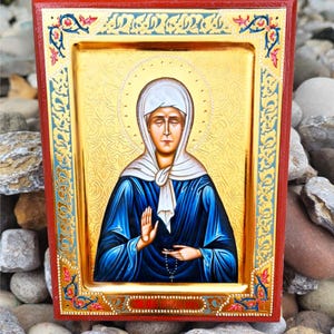 May include: A religious icon featuring a woman in a blue robe and white head covering, set against a gold background with a halo. The icon is framed in red with ornate gold and blue detailing, resting on a bed of stones.