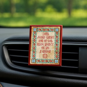 Könnte beinhalten: Ein rechteckiger Auto-Lufterfrischer mit rotem Rahmen und aufwendigem Design. Der Text lautet "Lord Jesus Christ Son of God Have Mercy On Me A Sinner" in roter Schrift. Der Hintergrund ist ein verschwommenes Grün.