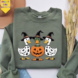 A green sweatshirt with a Halloween-themed graphic of three white ducks wearing witch hats and a carved pumpkin in the center.