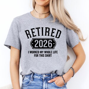 A woman wearing a grey t-shirt with the text "RETIRED 2026 I WORKED MY WHOLE LIFE FOR THIS SHIRT" printed on it.