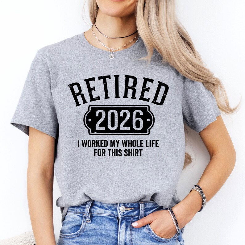 A woman wearing a grey t-shirt with the text "RETIRED 2026 I WORKED MY WHOLE LIFE FOR THIS SHIRT" printed on it.
