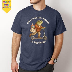 May include: Navy blue t-shirt featuring a cartoon frog riding a scooter, carrying a tote bag with groceries. The text reads "Off to help tiny humans do big things."