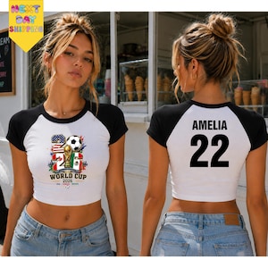 May include: A white and black raglan crop top featuring a 2026 World Cup graphic with a soccer ball and flags on the front. The back displays the name AMELIA and the number 22 in bold black text. The shirt has short black sleeves and a round neck.