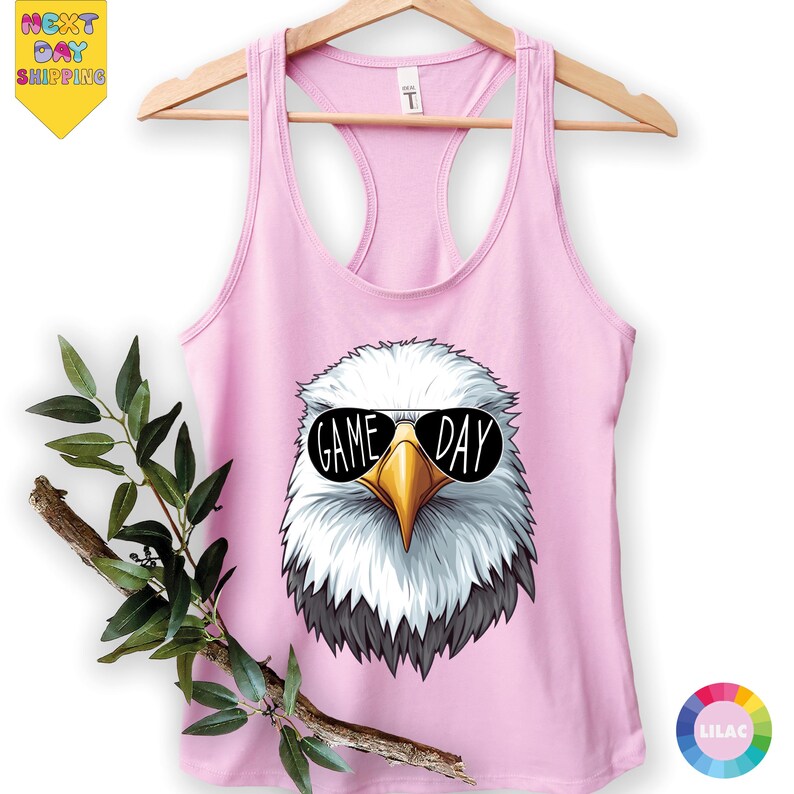 A pink tank top with a graphic of a white eagle wearing sunglasses and the text "GAME DAY" printed on it, hanging on a wooden hanger.