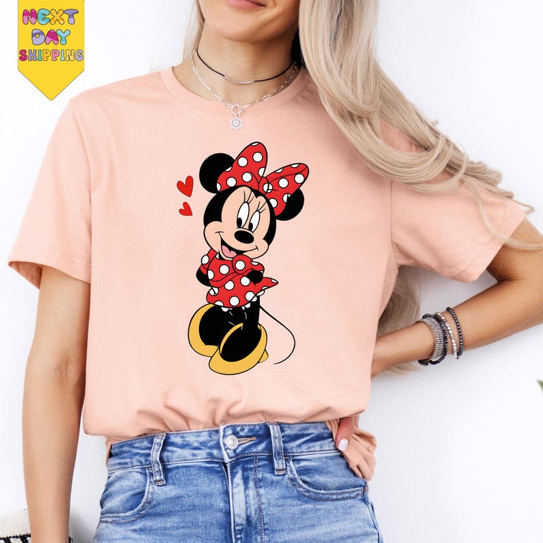 A woman wearing a pink t-shirt with a Minnie Mouse graphic and jeans, smiling and posing for the camera.