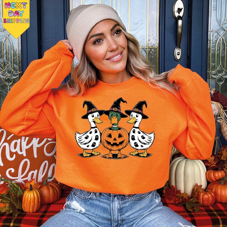A woman wearing an orange sweatshirt with a Halloween-themed graphic of three cartoon ducks and a jack-o-lantern.