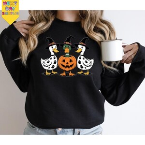 A woman is wearing a black sweatshirt with a Halloween-themed graphic design featuring three white ducks with black spots and a jack-o-lantern. She is holding a white mug in her hand.