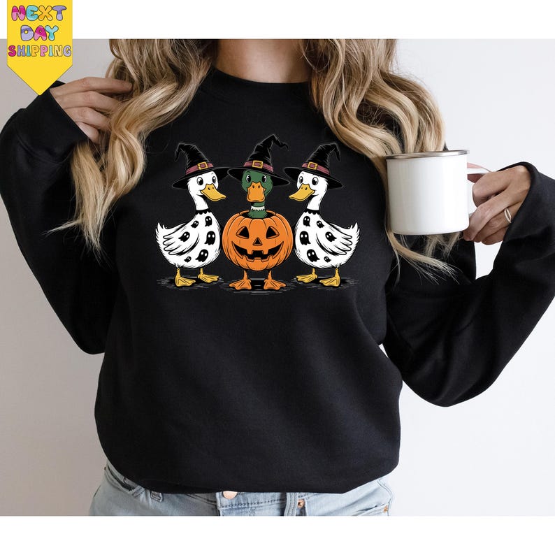 A woman is wearing a black sweatshirt with a Halloween-themed graphic design featuring three white ducks with black spots and a jack-o-lantern. She is holding a white mug in her hand.