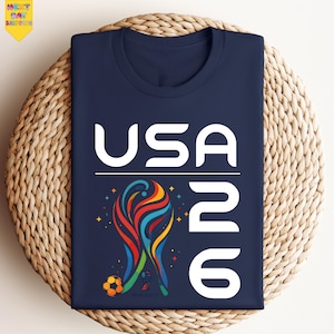May include: Navy blue t-shirt with the text "USA 26" in white. The shirt features a colorful graphic of a soccer ball and a stylized design. The shirt is folded on a woven surface.