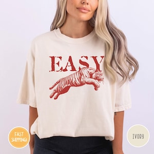 May include: Ivory-colored t-shirt with the word "EASY" in red block letters above a red tiger leaping across the front. The shirt has short sleeves and a relaxed fit.