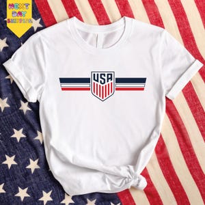 A white t-shirt with the text "USA" printed on it, along with a shield-shaped logo featuring red, white, and blue stripes. The t-shirt is placed on top of an American flag, which is spread out on a surface.
