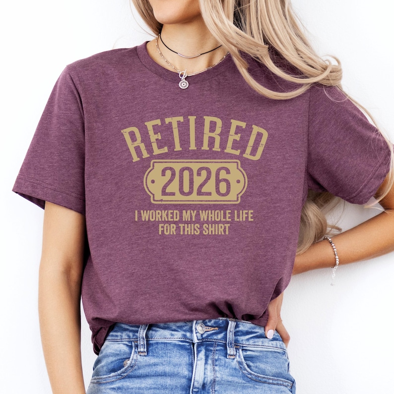 A woman wearing a purple t-shirt with the text "Retired 2026 I worked my whole life for this shirt" printed on it.