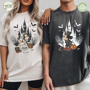 May include: A cream t-shirt featuring a graphic of a bride mouse in a wedding dress with a castle and bats, and a charcoal gray t-shirt featuring a graphic of a groom mouse in a top hat with a pumpkin, a castle, and bats in the background.