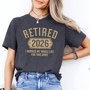 A woman wearing a gray t-shirt with the text "RETIRED 2026 I WORKED MY WHOLE LIFE FOR THIS SHIRT" printed on it.