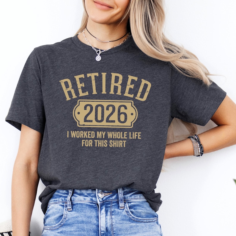 A woman wearing a gray t-shirt with the text "RETIRED 2026 I WORKED MY WHOLE LIFE FOR THIS SHIRT" printed on it.