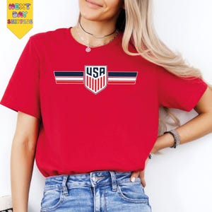 A woman wearing a red t-shirt with a graphic of the United States flag and blue jeans stands against a white background.