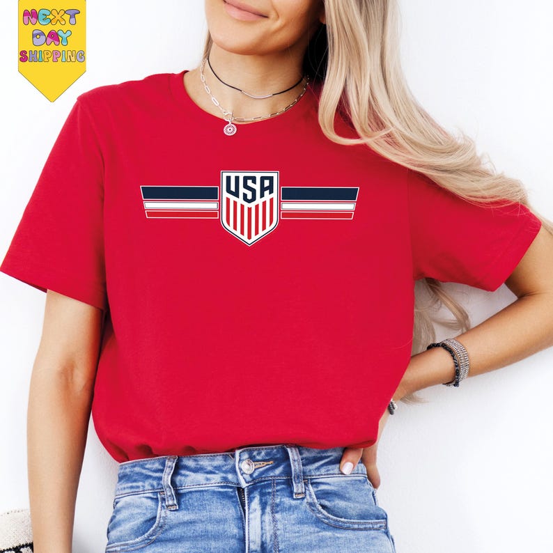A woman wearing a red t-shirt with a graphic of the United States flag and blue jeans stands against a white background.