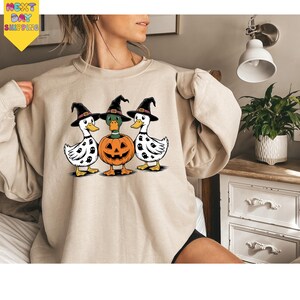 A woman is wearing a beige sweater with a Halloween-themed graphic of three ducks, one of which is holding a carved pumpkin.