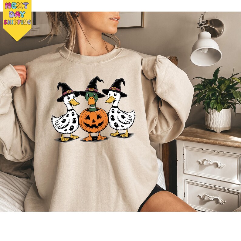 A woman is wearing a beige sweater with a Halloween-themed graphic of three ducks, one of which is holding a carved pumpkin.