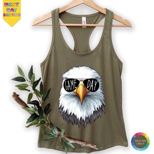 A green tank top with a graphic of a bald eagle wearing sunglasses and the text "GAME DAY" printed on it, hanging on a wooden hanger.