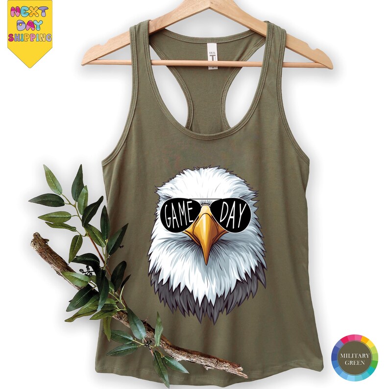 A green tank top with a graphic of a bald eagle wearing sunglasses and the text "GAME DAY" printed on it, hanging on a wooden hanger.