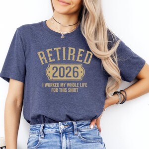 A woman wearing a blue t-shirt with the text "Retired 2026 I worked my whole life for this shirt" printed on it.