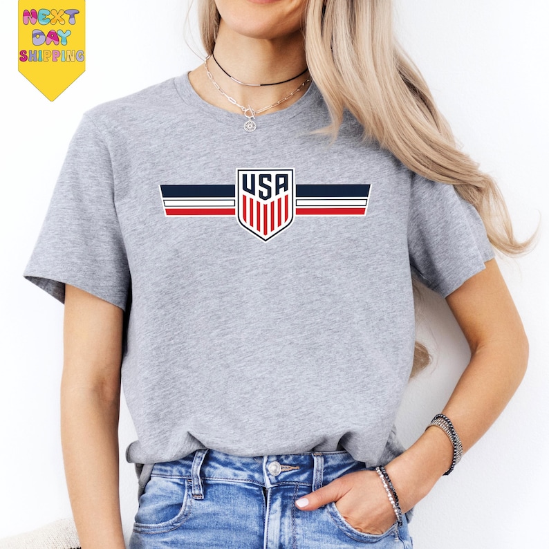 A woman wearing a gray t-shirt with a graphic of the United States flag and a shield design, paired with blue jeans.