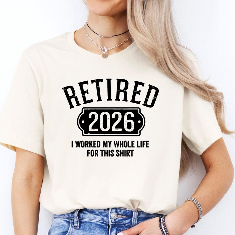 A woman wearing a white t-shirt with the text "RETIRED 2026 I WORKED MY WHOLE LIFE FOR THIS SHIRT" printed on it.