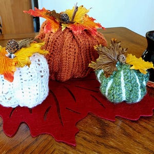 May include: A collection of handmade fall decorations, including crocheted pumpkins in white, orange, and green, adorned with faux leaves and pinecones. A dark brown mug and a golden pumpkin are also present, all arranged on a red leaf-shaped felt mat.