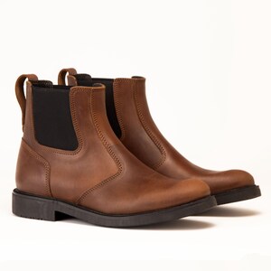 Pode incluir: Um par de botas Chelsea de couro castanho com pain&eacute;is laterais el&aacute;sticos pretos e puxadores. As botas t&ecirc;m uma sola baixa preta e um design cl&aacute;ssico. O couro tem uma apar&ecirc;ncia ligeiramente desgastada.