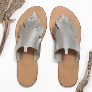 Handmade, top-grain, leather thong style sandal in metallic silver, with oak-leaf cutout design. Features deep brown colour, full-grain leather midsole. Classic, summer sandals perfect for beach, casual wear or vacation. Stylish and comfortable leather flip-flops with a natural feel. Lightweight and versatile for everyday use. Shown in an overhead view of the pair of sandals. Photographed on a white background with sand and driftwood, evoking a beachy, coastal vibe.