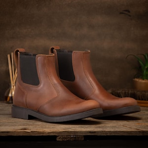 Pode incluir: Um par de botas Chelsea de couro castanho com pain&eacute;is laterais el&aacute;sticos. As botas t&ecirc;m um salto baixo e uma sola castanha escura. As botas s&atilde;o exibidas numa superf&iacute;cie de madeira.