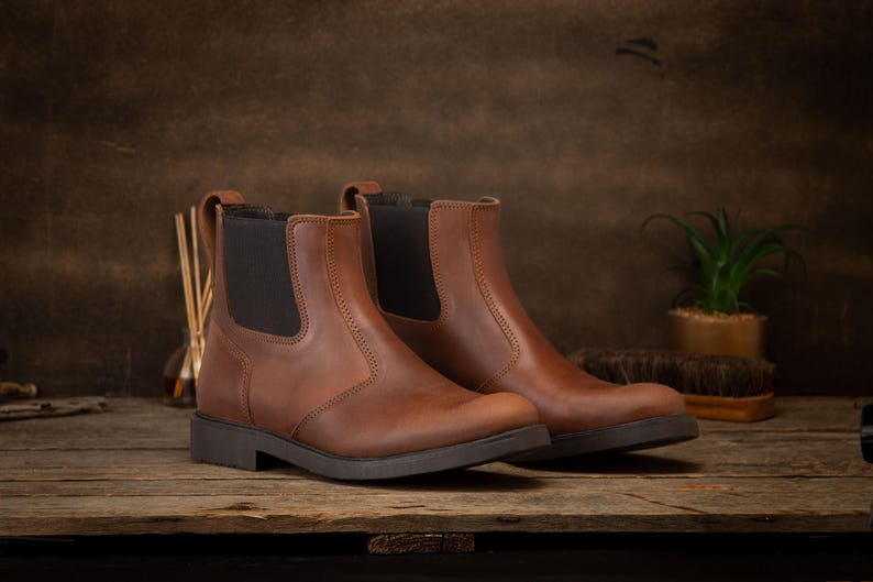 Pode incluir: Um par de botas Chelsea de couro castanho com pain&eacute;is laterais el&aacute;sticos. As botas t&ecirc;m um salto baixo e uma sola castanha escura. As botas s&atilde;o exibidas numa superf&iacute;cie de madeira.