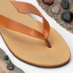 May include: A pair of tan leather thong sandals with a simple design. The sandals are made of a single piece of leather, with a strap that goes over the toes and a thong that goes between the toes.
