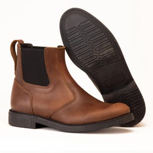 Pode incluir: Um par de botas Chelsea de couro castanho. Uma bota est&aacute; em p&eacute;, mostrando o painel lateral el&aacute;stico e a presilha. A outra bota est&aacute; angulada, revelando a sola de borracha preta. As botas t&ecirc;m um design cl&aacute;ssico e robusto.