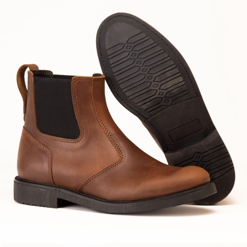 Pode incluir: Um par de botas Chelsea de couro castanho. Uma bota est&aacute; em p&eacute;, mostrando o painel lateral el&aacute;stico e a presilha. A outra bota est&aacute; angulada, revelando a sola de borracha preta. As botas t&ecirc;m um design cl&aacute;ssico e robusto.