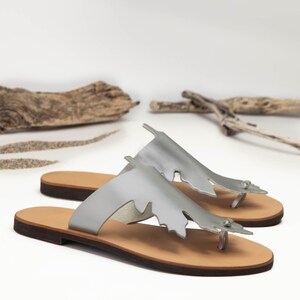 Handmade, top-grain, leather thong style sandal in metallic silver, with oak-leaf cutout design. Features natural hide  color, full-grain leather midsole. Classic, summer sandals perfect for beach, casual wear or vacation. Stylish and comfortable leather flip-flops with a natural feel. Lightweight and versatile for everyday use. Shown in an angled view of the pair of sandals. Photographed on a white background with sand and driftwood, evoking a beachy, coastal vibe.
