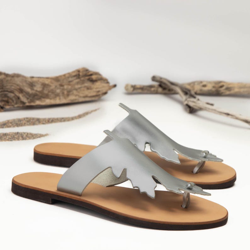 Handmade, top-grain, leather thong style sandal in metallic silver, with oak-leaf cutout design. Features natural hide  color, full-grain leather midsole. Classic, summer sandals perfect for beach, casual wear or vacation. Stylish and comfortable leather flip-flops with a natural feel. Lightweight and versatile for everyday use. Shown in an angled view of the pair of sandals. Photographed on a white background with sand and driftwood, evoking a beachy, coastal vibe.