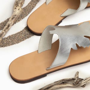 Handmade, top-grain, leather thong style sandal in metallic silver color, with oak-leaf cutout design. Features natural hide colour, full-grain leather midsole. Classic, summer sandals perfect for beach, casual wear or vacation. Stylish and comfortable leather flip-flops with a natural feel. Lightweight and versatile for everyday use. Shown in an overhead angled rear view of the pair of sandals. Photographed on a white background with sand and driftwood, evoking a beachy, coastal vibe.