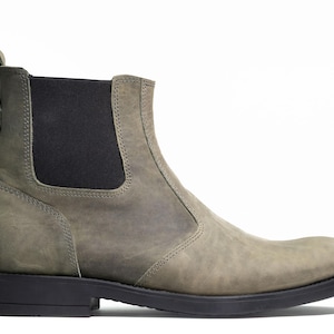 Pode incluir: Vista lateral de uma bota Chelsea cinzenta com sola preta e painel lateral el&aacute;stico. A bota tem uma aba na parte de tr&aacute;s e &eacute; feita de um material texturizado semelhante ao couro. A bota tem aproximadamente 15 cm de altura.